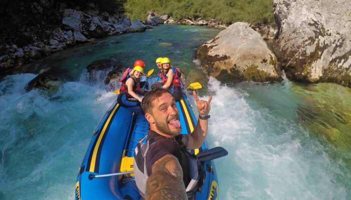 Soča Rafting Selfie Guide and smiling guests take selfie on Soča River rafting tour, sunny summer day with rocks in background.