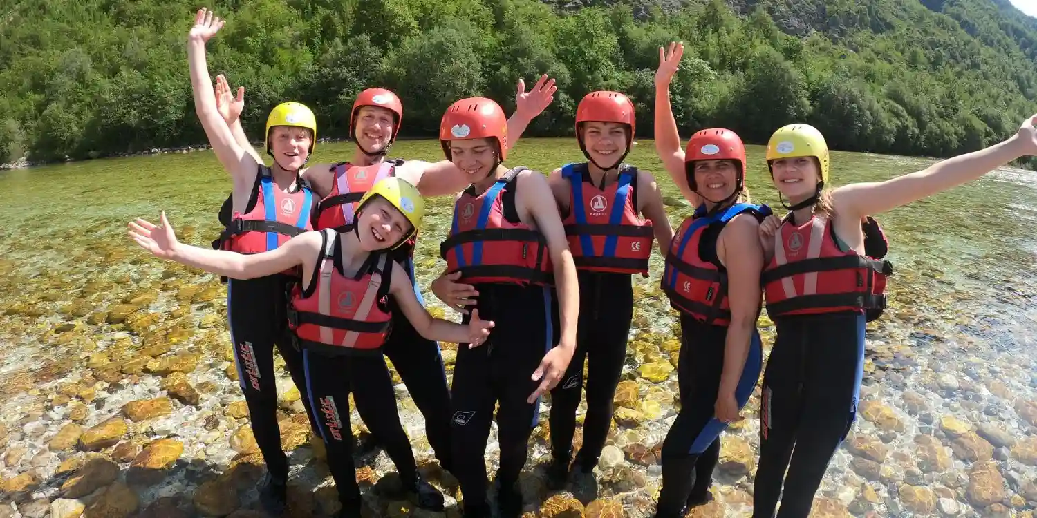 Happy family with teenagers wearing full rafting safety gear, including helmets and life jackets, standing in the emerald Soča River in Bovec, Slovenia, during a sunny whitewater rafting trip.