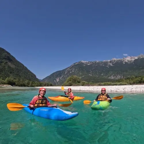 Smiling guests paddling their kayaks on a calm, crystal-clear section of the Soča river with breathtaking mountain peaks in the background.