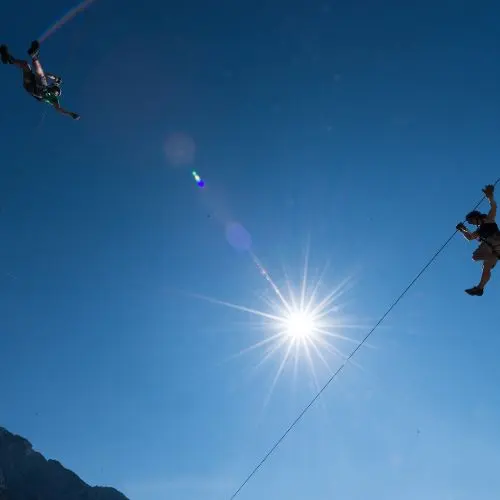 An adventurous guest ziplining high above the ground with a beautiful sun flare and Julian Alps in the distance.