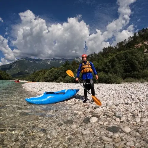 A guest standing on a pebble beach by the Soča river, fully equipped in kayak gear, holding a paddle with a kayak on the ground.