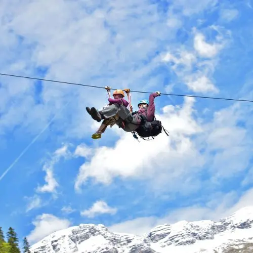 A guest and a professional guide flying together in a tandem zipline position over the mountain landscape.