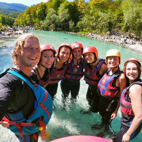 A happy group of friends and their guide standing in the crystal clear Soča river during a bachelorette party rafting trip in Bovec, Slovenia.