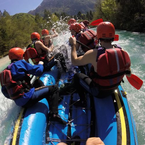 Un gruppo di amici con caschi rossi e gialli pagaia insieme su un gommone blu tra le rapide del fiume Isonzo a Bovec, in Slovenia.